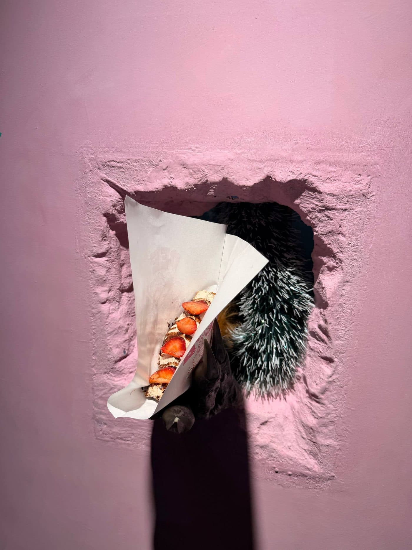 A spiky green paw pushing a strawberry Fried Sando out through the hole in the pink wall